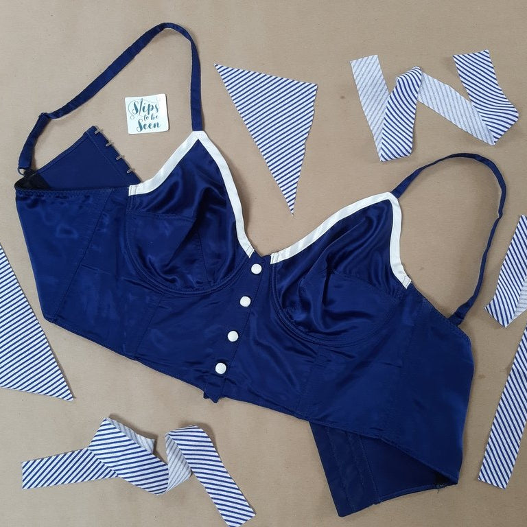 The image shows a 8 Huit blue corset with with white buttons, adjustable straps and soft cups. There is a brown background, the Slips to be Seen logo and strips of blue and white fabric.